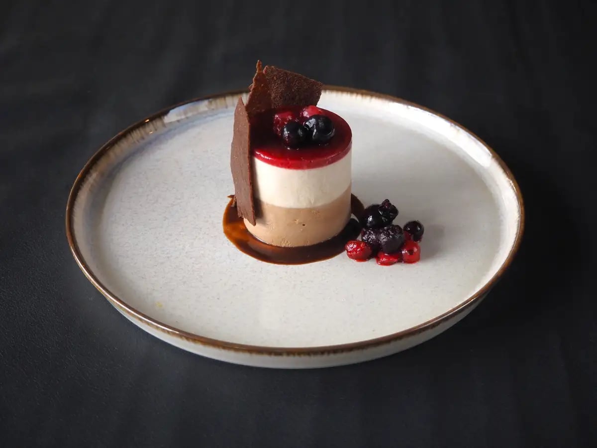 White Chocolate and Bitter Chocolate W Mousse with Raspberry Sauce. Layers of white and bitter chocolate mousse are topped with deep red raspberry sauce and berries. Decorated with a chocolate tuile, offering a contrast between smooth mousse and crispy, light texture—perfect for Christmas.