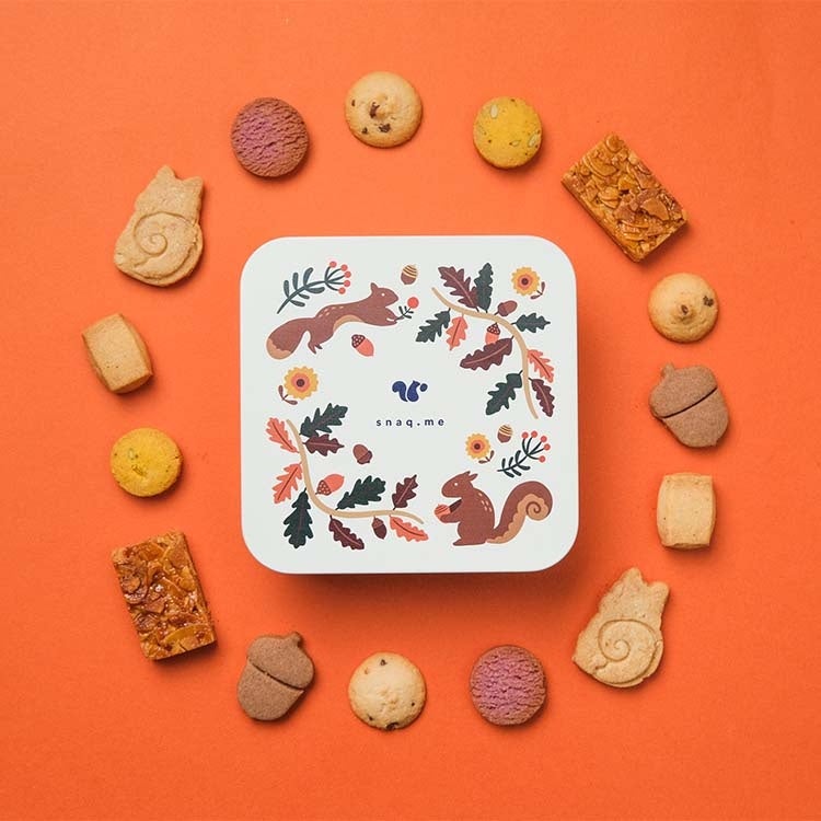 This is a cozy cookie tin inspired by autumn forests, featuring acorns and squirrels. It contains a variety of treats like almond florentines, purple sweet potato cookies, and pumpkin cookies, each representing the harvest season. The cookies are crafted to highlight natural flavors with a mix of crispy, crumbly, crunchy, and firm textures, all in one charming tin. The design evokes a warm autumn forest scene with colorful leaves, nuts, and flowers, perfect for a relaxing snack time.